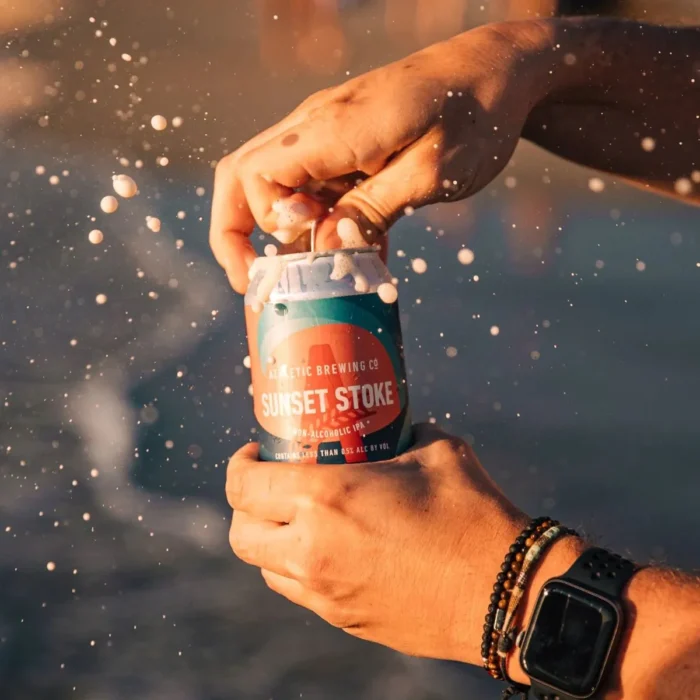 Sunset Stride non-alcoholic beer can on a beach with ocean in the background.