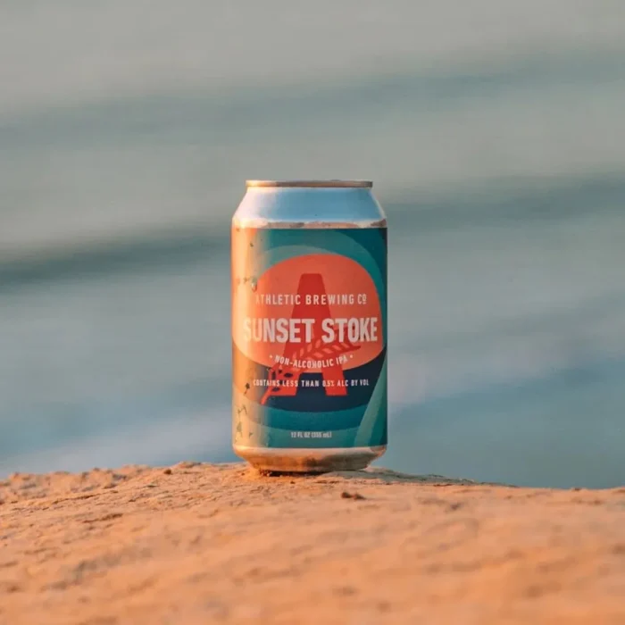 Sunset Stride non-alcoholic beer can on a beach with ocean in the background.