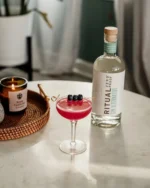 Ritual Zero Proof bottle with a vibrant red cocktail garnished with blueberries on a cozy table setup.