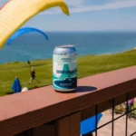 Can of Athletic Brewing Co.'s non-alcoholic beer on a wooden railing overlooking a scenic coastline with paragliders in the background.