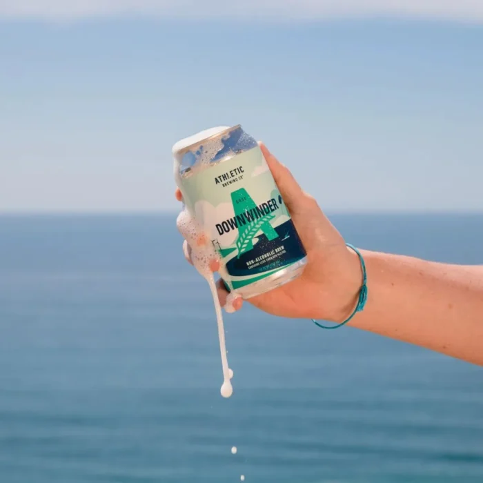 Hand holding a frothy can of Athletic Brewing Co.'s non-alcoholic beer by the ocean, perfect for refreshing seaside enjoyment.