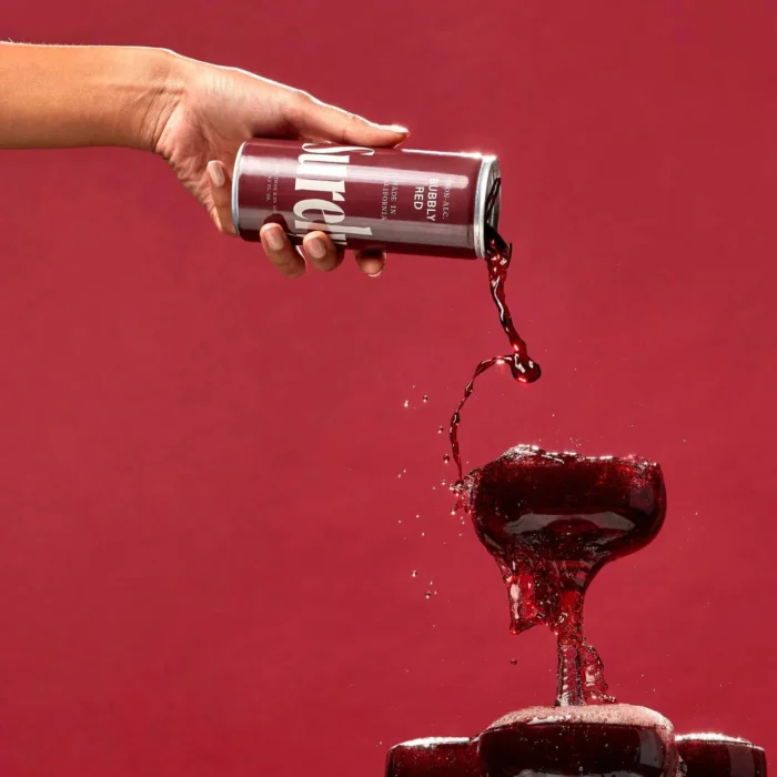 Pouring a rich red beverage from a can into stacked glasses against a bold red background.