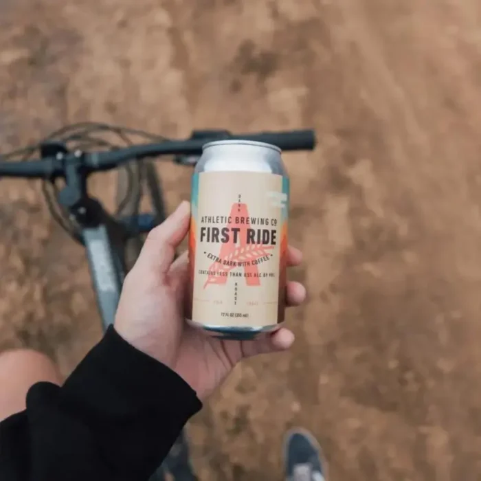 Hand holding a can of First Ride non-alcoholic coffee-infused beer on a dirt trail, showcasing an adventurous and refreshing beverage choice.