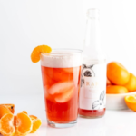 Rare Brew served in a glass with ice and topped with a slice of mandarin orange, accompanied by a bottle of Rare Brew and fresh citrus fruits in the background.