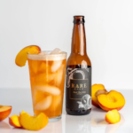 A glass filled with golden, iced peach-flavored beverage garnished with a peach slice, paired with a bottle labeled 'Rare Brew: Peach Tree Hops,' surrounded by fresh peach slices.