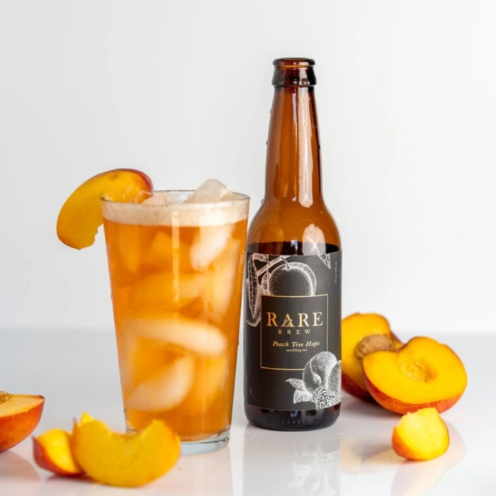 A glass filled with golden, iced peach-flavored beverage garnished with a peach slice, paired with a bottle labeled 'Rare Brew: Peach Tree Hops,' surrounded by fresh peach slices.