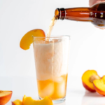 Pouring golden liquid into a glass filled with creamy foam and ice, garnished with a peach slice, surrounded by fresh peach pieces.