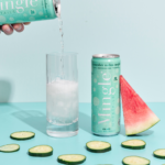 Cucumber Melon Mojito Mocktail being poured into a tall glass filled with ice, displayed alongside fresh cucumber slices and a watermelon wedge on a pastel blue background.