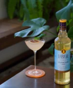Liquor bottle beside a cocktail glass on a wooden surface with greenery in the background.