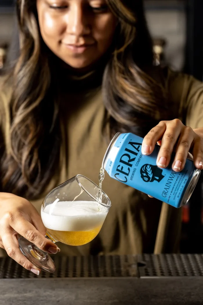 Person pouring Ceria Brewing Grainwave non-alcoholic beer into a glass, highlighting golden color and foam.