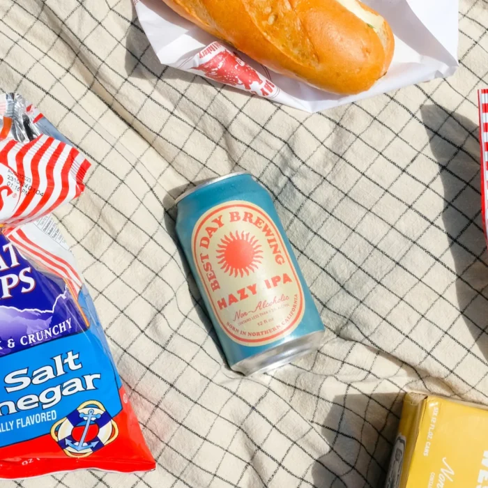 Picnic setup featuring a can of non-alcoholic Hazy IPA, surrounded by snacks like chips, a sandwich, and cheese on a checkered blanket in the sun.