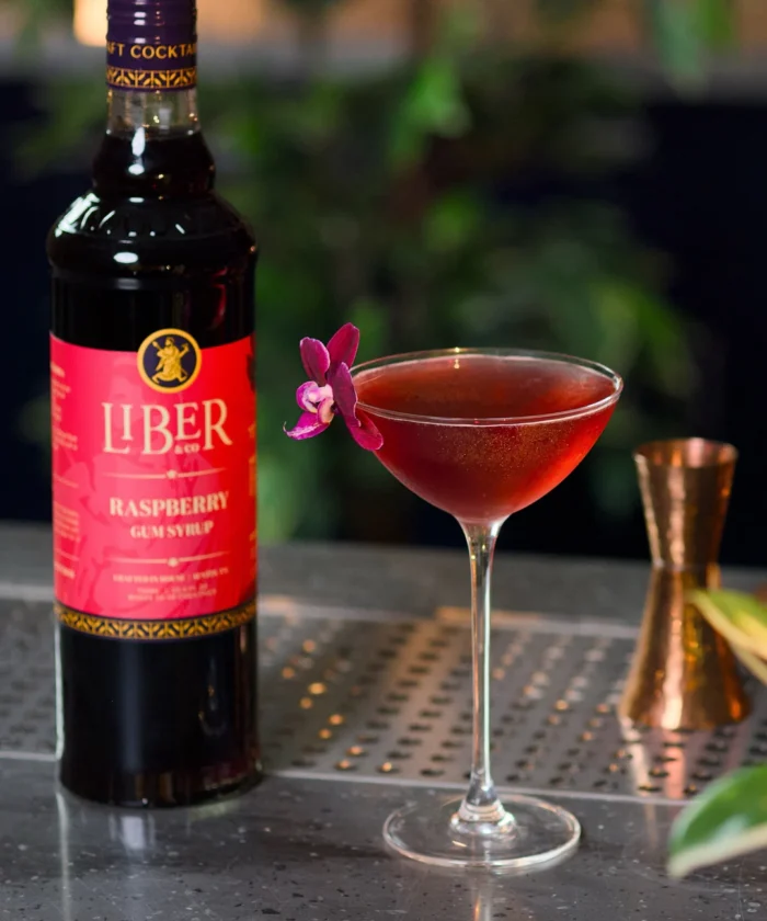 ark liquor bottle beside a red cocktail in a glass with a flower garnish on a bar counter.