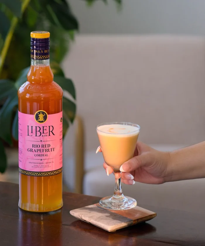 Orange liquor bottle next to a frothy cocktail in a glass held by a hand.