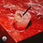 Vibrant red cocktail with a lime garnish and straw on a textured red marble surface.
