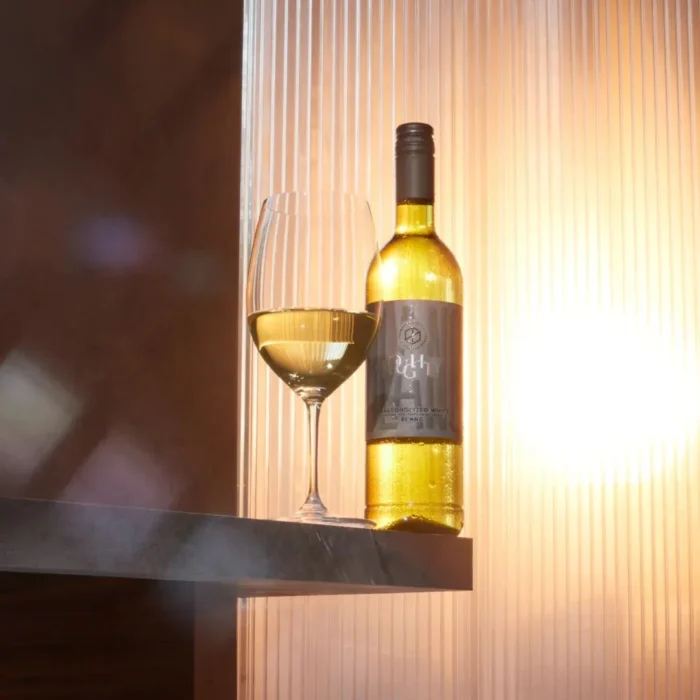 Bottle of white wine with a filled glass on a shelf, illuminated by warm, golden lighting.