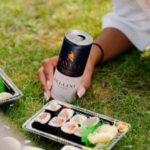 Hand holding a Savile can near sushi trays on a picnic setup over grass.