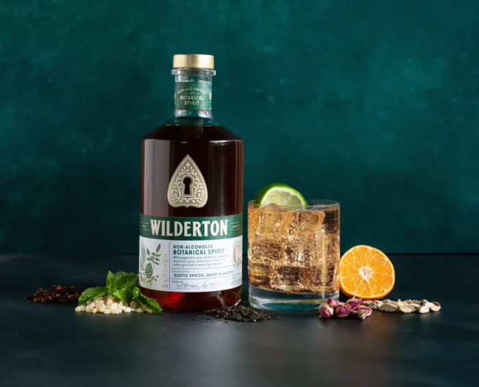 Wilderton non-alcoholic spirit bottle with a lime-garnished cocktail, herbs, and citrus on a dark backdrop.