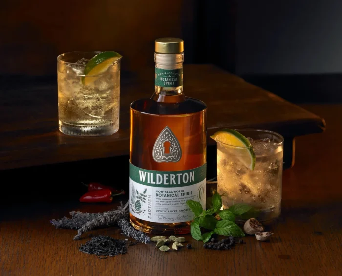 Wilderton non-alcoholic amber spirit bottle with herb and spice garnished cocktails on a dark table setting.