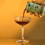 Little Saints can pouring a dark liquid into a coupe glass against a warm orange background.