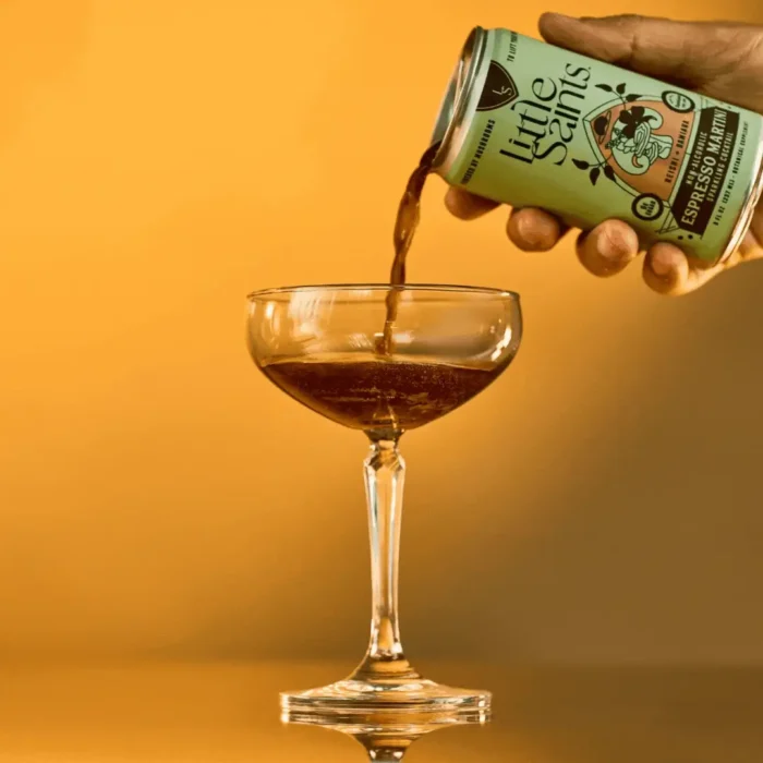 Little Saints can pouring a dark liquid into a coupe glass against a warm orange background.