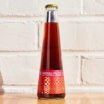 Amaro Falso non-alcoholic cocktail bottle with a vibrant red label against a white brick background.