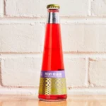 Vivid red non-alcoholic aperitif bottle with purple and gold accents against a white brick background.