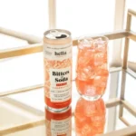 Hella Bitters & Soda can with a glass of sparkling pink beverage and ice on a reflective surface.