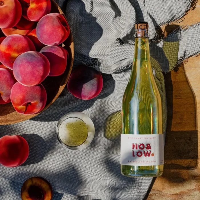 No & Low sparkling peach wine bottle with a bowl of ripe peaches and a glass in natural sunlight.