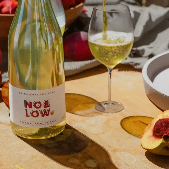 No & Low sparkling peach wine bottle with a glass being poured and fresh peaches on a sunny table.