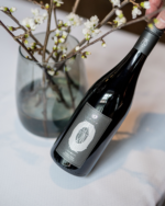 Elegant bottle of red wine placed beside a glass vase with blooming branches on a light surface.