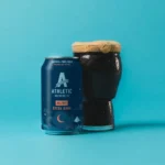 Can of Athletic Brewing Co.'s All Out Extra Dark non-alcoholic beer next to a glass of rich, dark brew with a frothy head against a bright blue background.