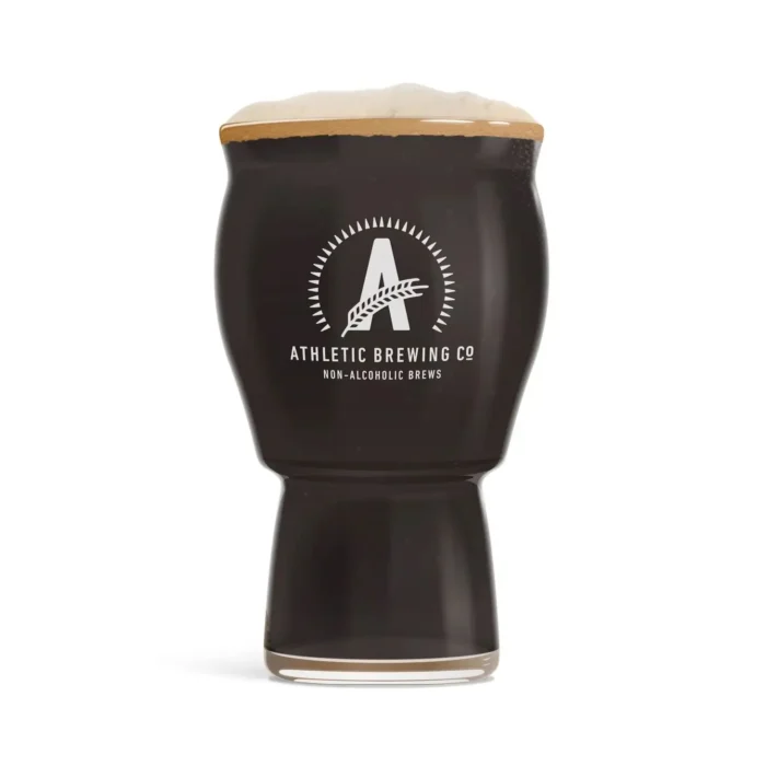 Glass of Athletic Brewing Co.'s All Out Extra Dark non-alcoholic beer, showcasing a rich, dark color and frothy head with branded logo on the glass.