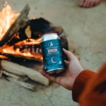 Hand holding a can of Athletic Brewing Co.'s All Out non-alcoholic extra dark beer by a campfire, perfect for cozy outdoor moments.