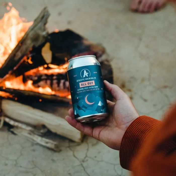 Hand holding a can of Athletic Brewing Co.'s All Out non-alcoholic extra dark beer by a campfire, perfect for cozy outdoor moments.