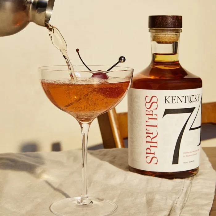 piritless Kentucky 74 non-alcoholic bourbon bottle beside a freshly poured cocktail garnished with a cherry, set in a warm, sunlit atmosphere.