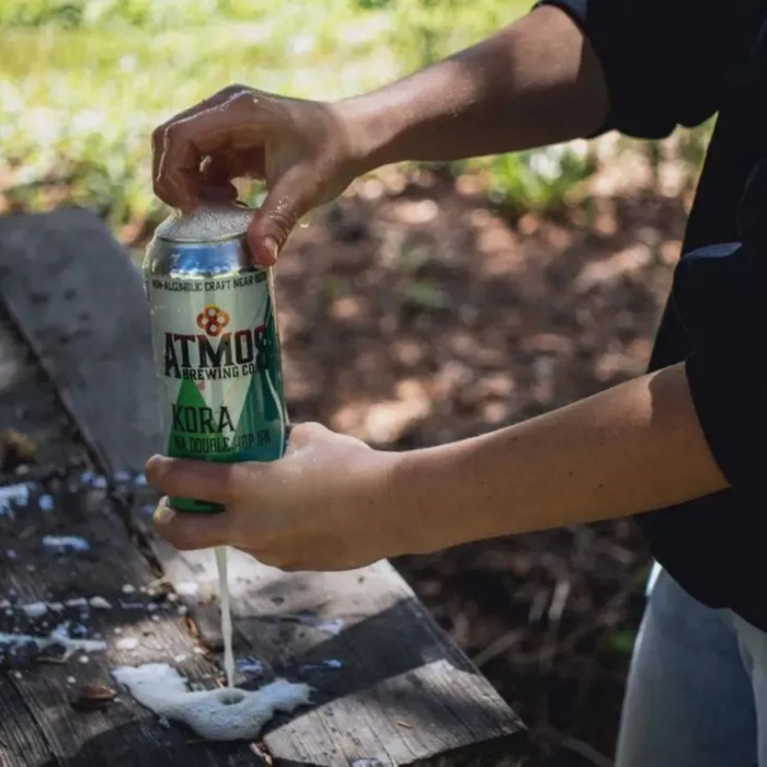 Opening a can of Atmos Brewing Co. Kora NA Double Hop IPA outdoors, with frothy foam spilling over, capturing a refreshing moment in nature.