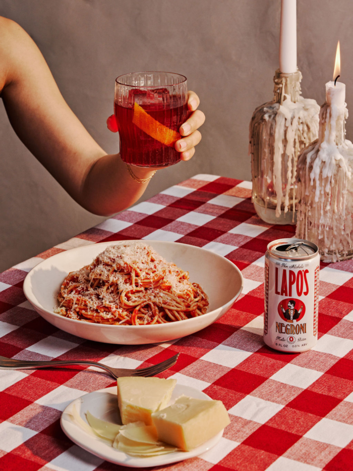 Plate of spaghetti with Lapo's Negroni can and red cocktail on a checkered table with candles.