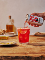 Pouring a red cocktail into a glass with Lapo canned drink on a wooden table with snacks and bottle.