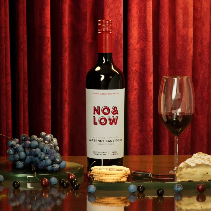 Bottle of No & Low alcohol-removed Cabernet Sauvignon with a glass, grapes, cheese, and crackers, set against a red curtain backdrop.