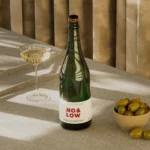 No & Low sparkling wine bottle with a glass and a bowl of olives on a sunlit table.