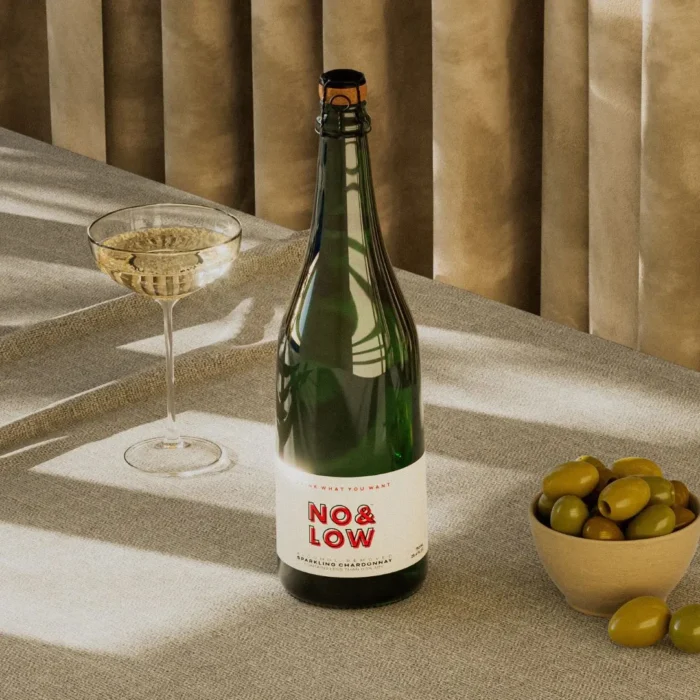 No & Low sparkling wine bottle with a glass and a bowl of olives on a sunlit table.