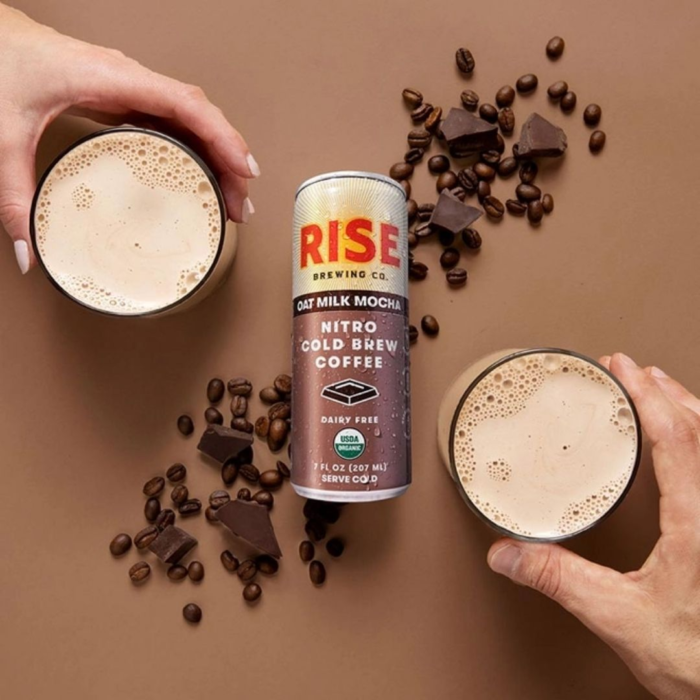 Rise Nitro Oat Milk Mocha can with two glasses, coffee beans, and chocolate pieces on a brown background.