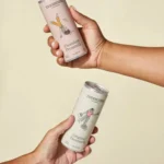 Two hands holding Treehouse Naturals Pecanmilk cans, one Chocolate Pecanmilk and one Original Pecanmilk, against a beige background.