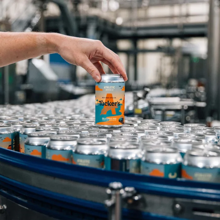 Production line showcasing Athletic Brewing Co.'s Tucker's West Coast Style IPA cans, with a hand selecting one, highlighting quality control and craftsmanship.