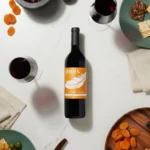 Flat lay of a wine bottle surrounded by glasses, dried fruits, olives, and cheese on a white table.