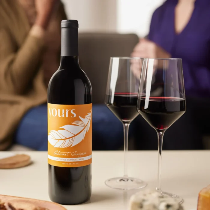 Bottle of non-alcoholic red wine with two glasses, set in a cozy setting with friends chatting in the background.