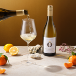Bottle of white wine being poured into a glass, surrounded by fresh citrus fruits and floral accents on a warm-toned table.