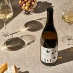 Bottle of Sovi Chenin Blanc with glasses of white wine, cheese, and olives on a sunlit linen table.