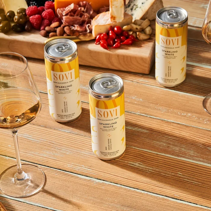 Sovi Sparkling White non-alcoholic wine cans on a wooden table with charcuterie board and glasses of wine.
