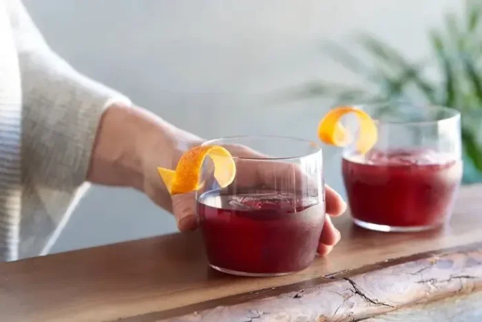 Hand holding a glass of red non-alcoholic cocktail garnished with orange peel, perfect for gatherings.